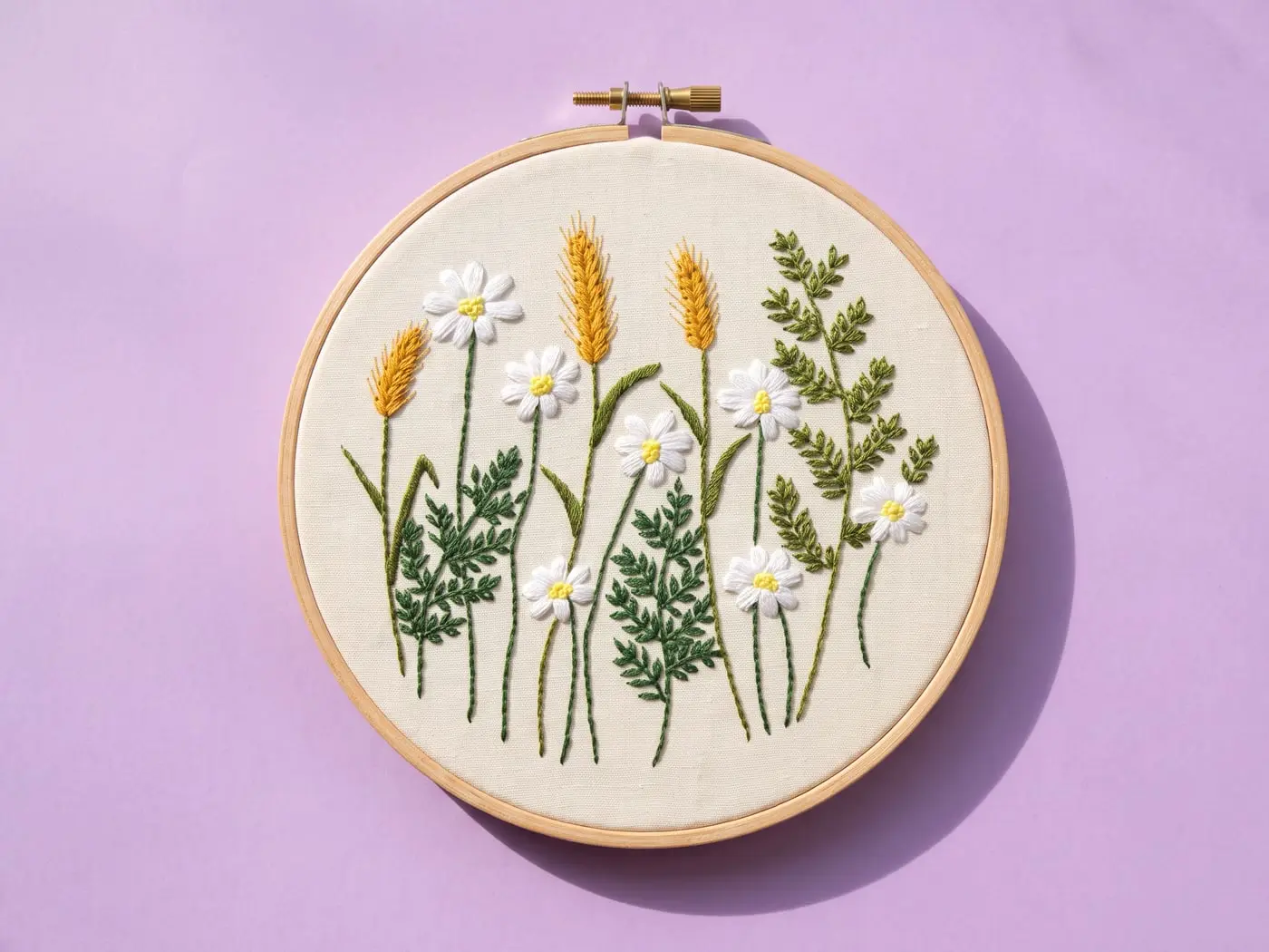Tambour brodé d'une prairie de marguerites et d'épis de blé, posé sur un papier lilas