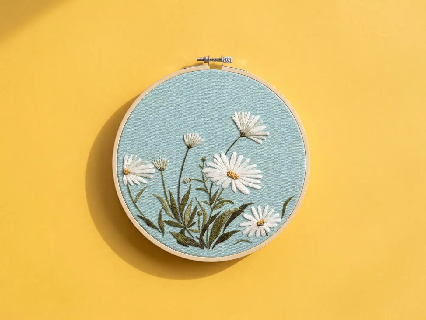 Tambour brodé de marguerites blanches sur une toile bleu-vert, posé sur un papier mimosa