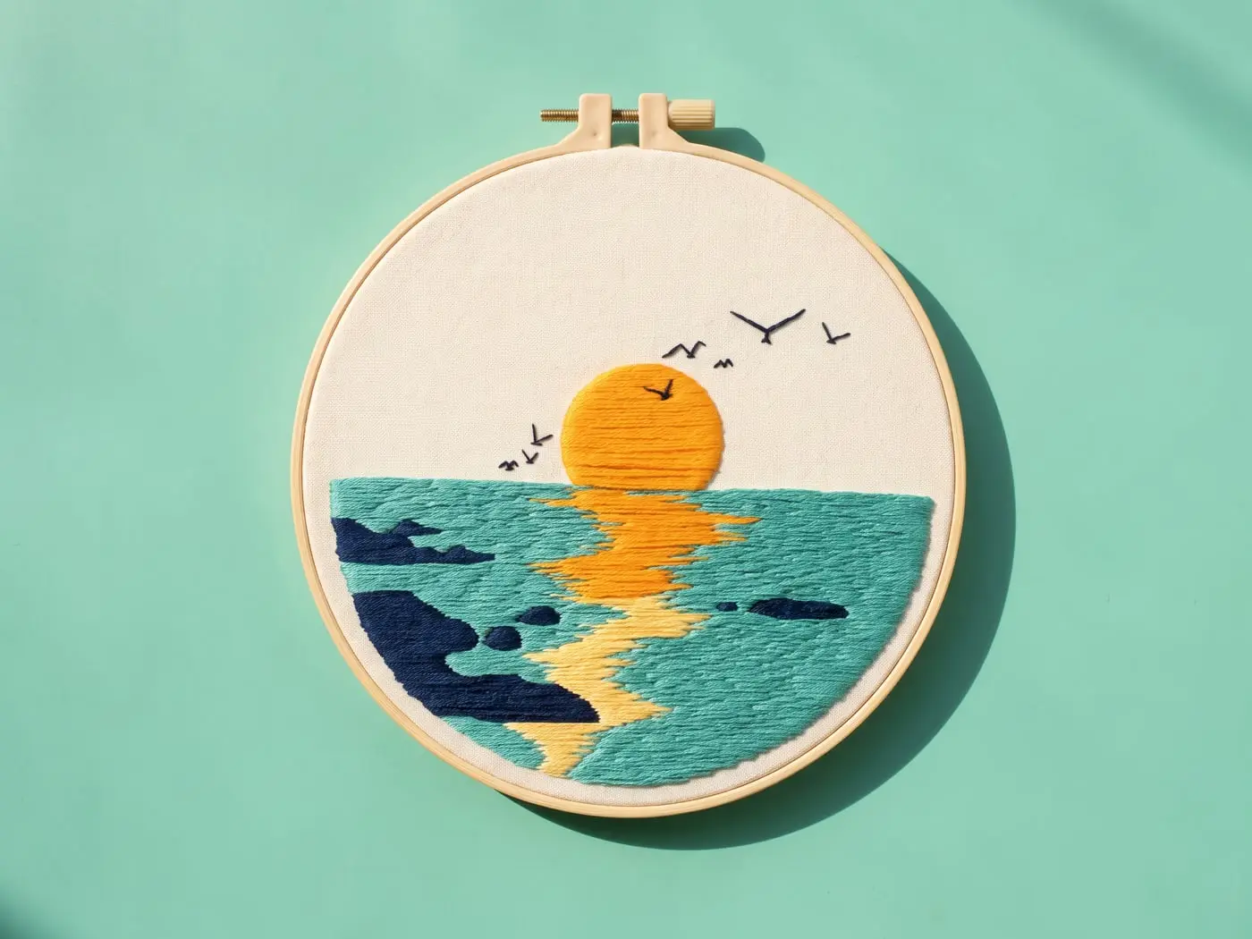 Tambour brodé d'un soleil orange couchant sur une mer turquoise, posé sur un papier menthe